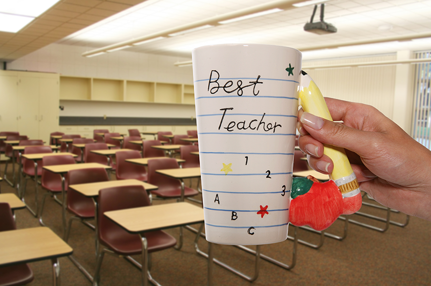 a teacher holds up a mug in front of an empty classroom that reads: "Best Teacher"