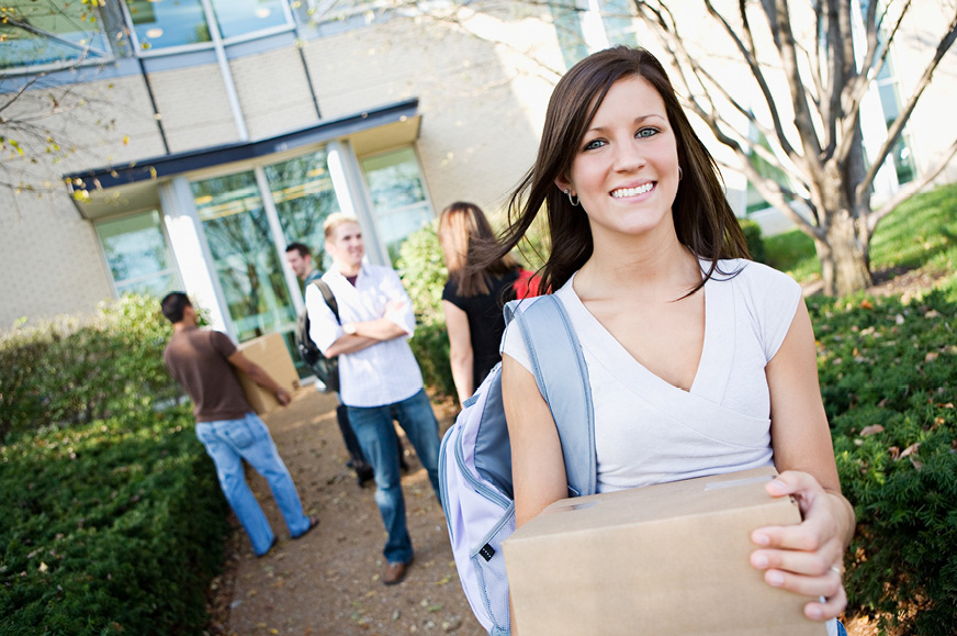 College-aged girl carrying box in front of apartment building