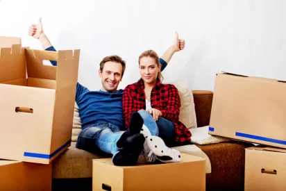a couple sits on a couch with empty moving boxes, the man has two big thumbs up in triumph