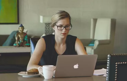 a woman with glasses looks at a mac laptop with a cup of coffee and a muffin