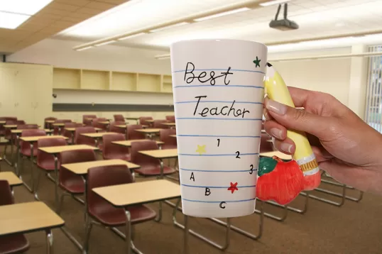 a teacher holds up a mug in front of an empty classroom that reads: "Best Teacher"